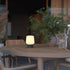 Lysende trådløs bordlampe fra sackit på et rundt trebord i en lun spisestue med grønne planter og glass i bakgrunnen