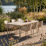 Kirra spisebord med seks lyse hagestoler dekket for måltid på en solrik terrasse ved innsjøen omgitt av frodige planter fra sackit