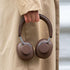 Brune hodetelefoner med over ear design holdes i hånden foran en beige frakk med polstrede øreputer og buet hodebøyle