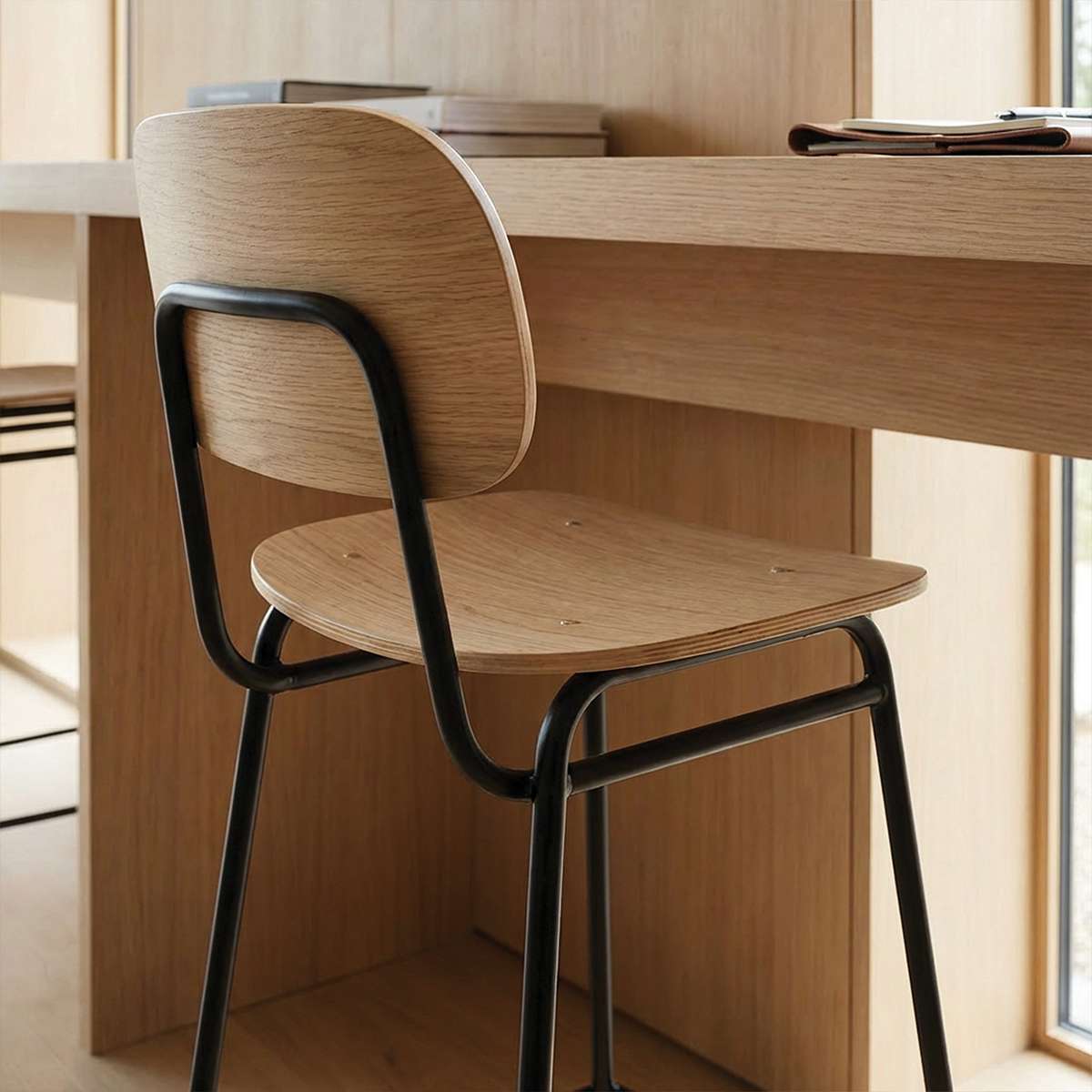 Barkrakk i lyst tre med buet ryggstøtte og svart understell ved et skrivebord i tre. Et rent og industrielt uttrykk for moderne innendørs bruk