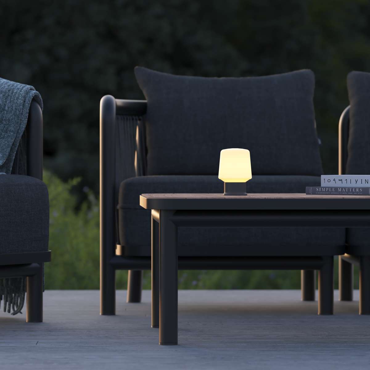 Liten trådløs bordlampe med varmt lys på et mørkt utebord mellom elegante loungestoler og sofaer i en nordisk utendørssetting