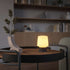 Trådløs bordlampe fra sackit med varmt lys på et rundt trebord i en stilfull nordisk stue med hyller og dekorative detaljer