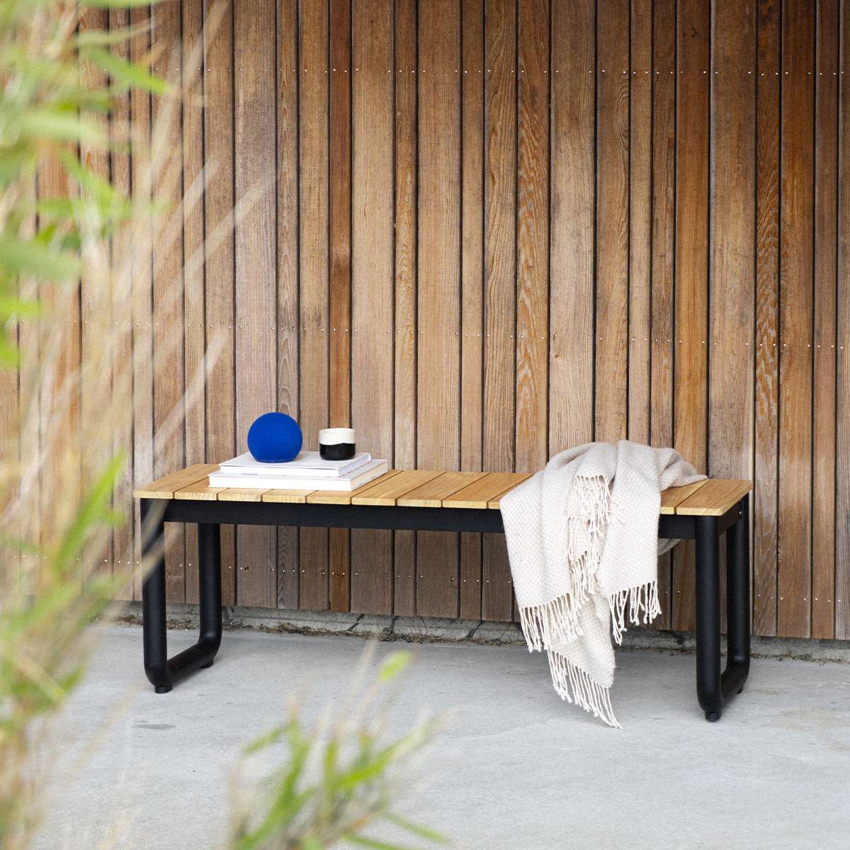 sackit utendørs benk i teak og sort pulverlakkert aluminium mot en trekledd vegg med pledd og dekorative detaljer setter en lun nordisk stemning