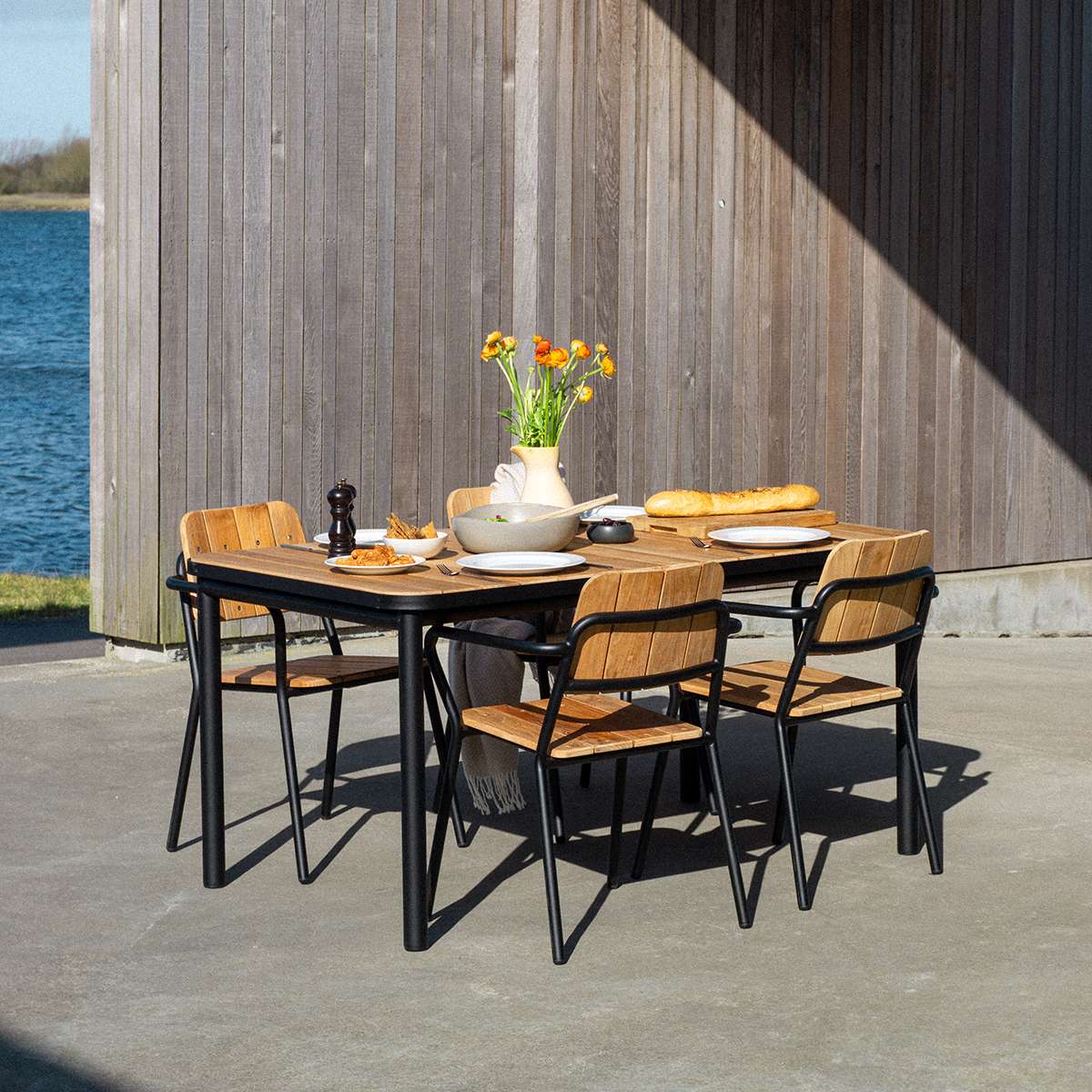 Kirra Dining Table 150x90 i pulverlakkert aluminium med ubehandlet teakplate i naturlig utendørsmiljø.