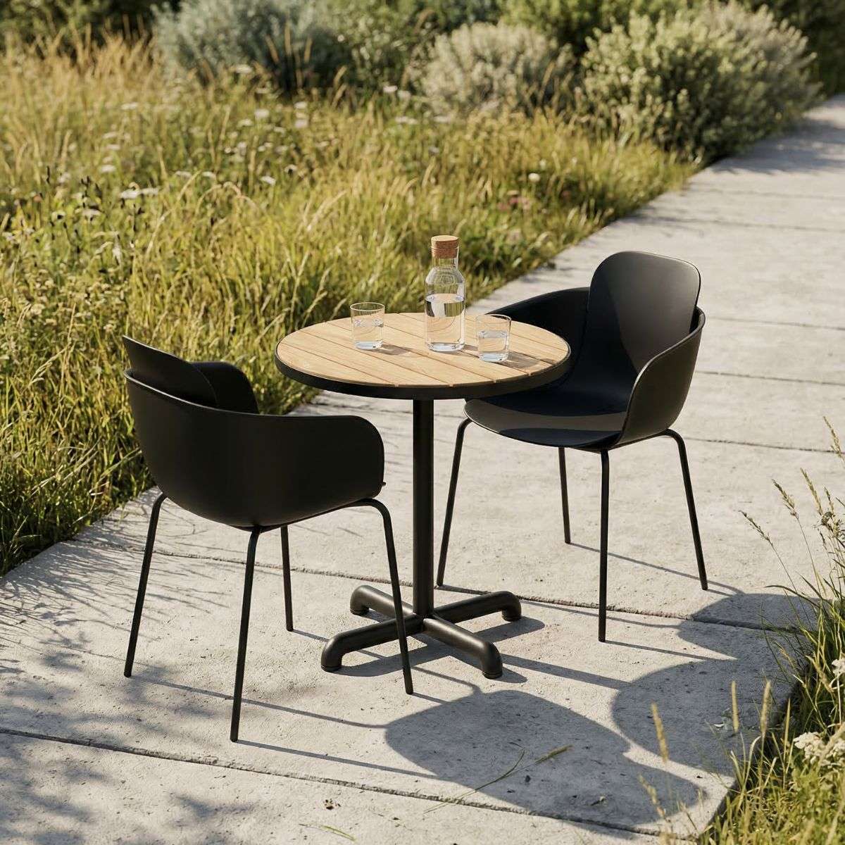Rundt utebord i teak med to sorte hagestoler fra sackit på en solrik terrasse omgitt av grønne planter og høyt gress