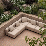 Beige femseters modulbasert hjørnesofa fra sackit på en teglbelagt terrasse omgitt av blomstrende planter og grønne busker