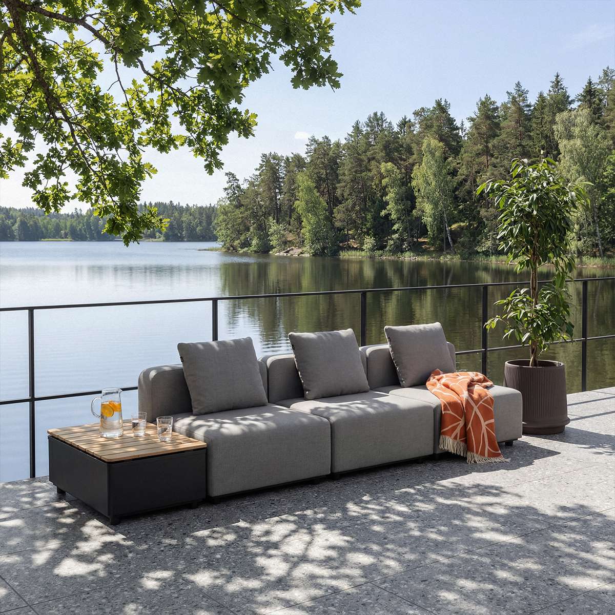 Modulbasert grå utesofa med puter og oransje pledd på terrasse ved innsjøen designet i Danmark av sackit Norway for nordisk vær