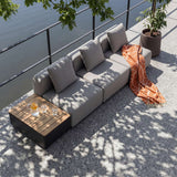 Grå Cobana loungesofa med tre sitteplasser og teakbord med drikke på solrik terrasse under grønne trær fra sackit