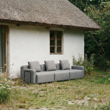 sackit Cobana Lounge Sofa i grå tekstil med tre sitteplasser foran en stråtakt hytte omgitt av grønt gress og frodig natur