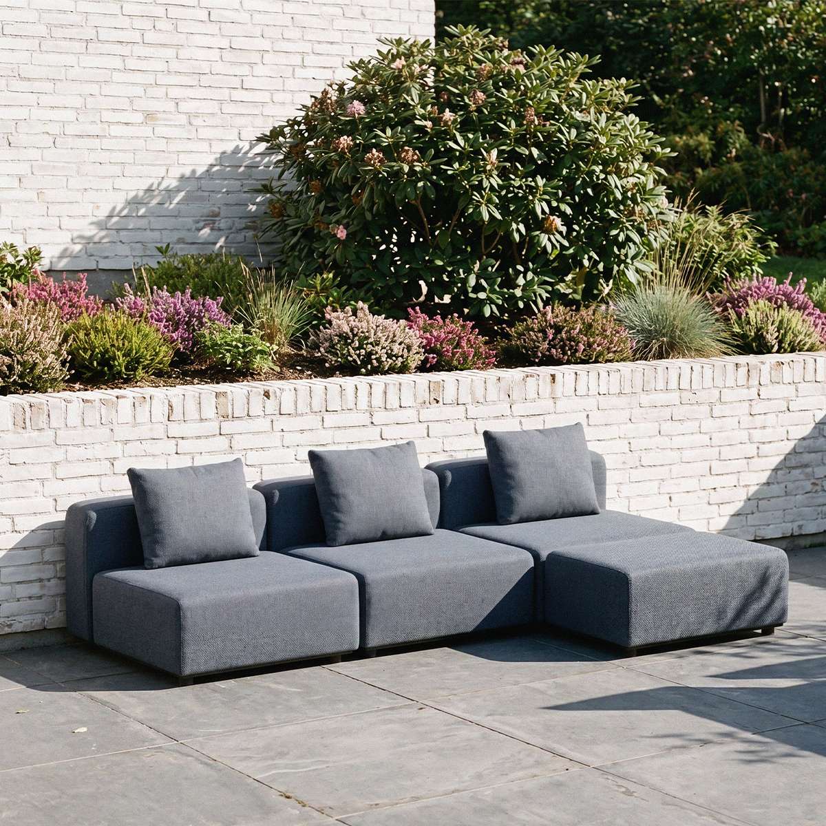 Mørkegrå modulbasert utesofa med tre sitteplasser og løse ryggputer på en solrik terrasse omgitt av grønne planter og blomstrende bed fra sackit