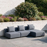 Mørkegrå modulbasert utesofa med tre sitteplasser og løse ryggputer på en solrik terrasse omgitt av grønne planter og blomstrende bed fra sackit
