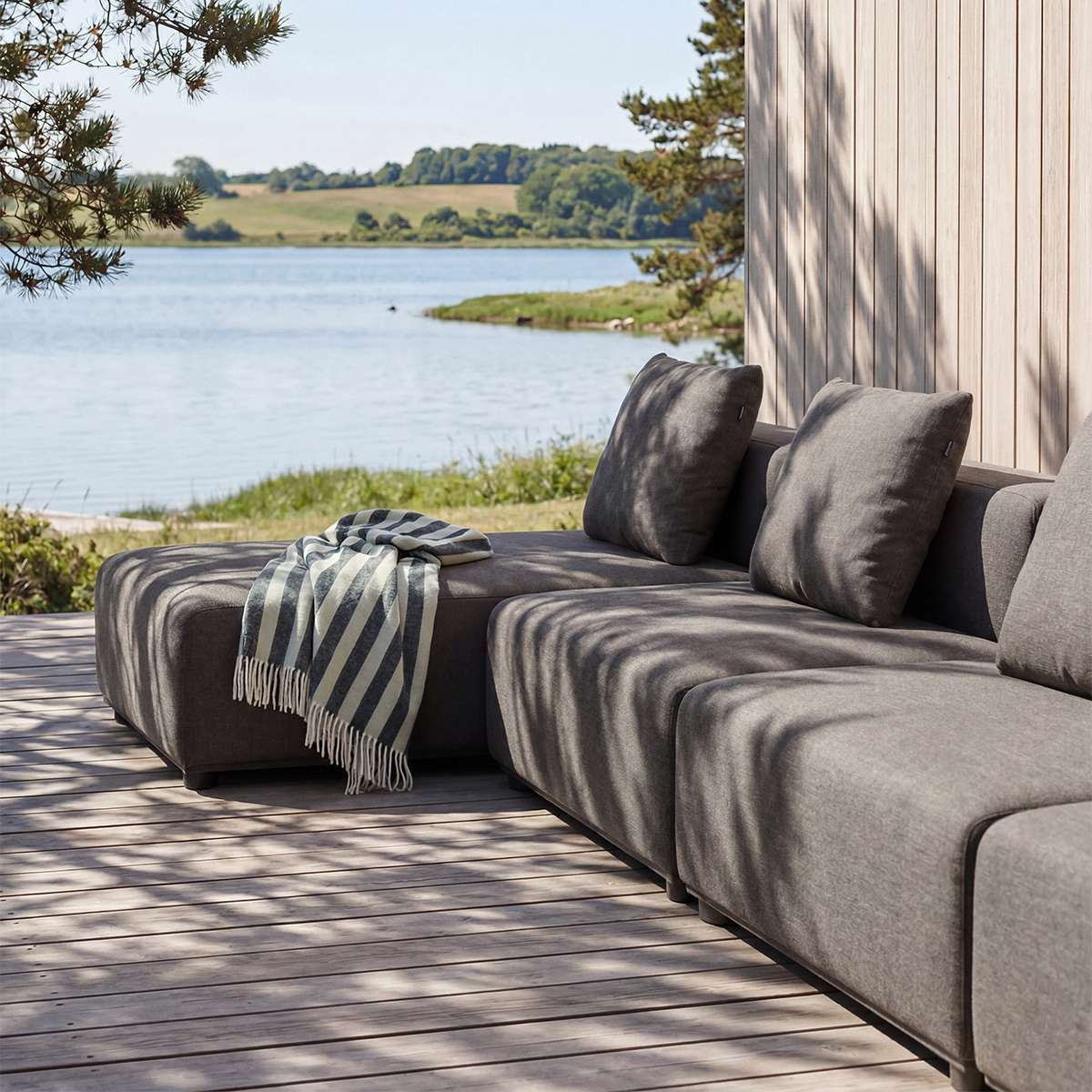Mørk grå modulsofa med sjeselong fra sackit Norway på treterrasse ved vannet med puter og stripete pledd i nordisk utemiljø