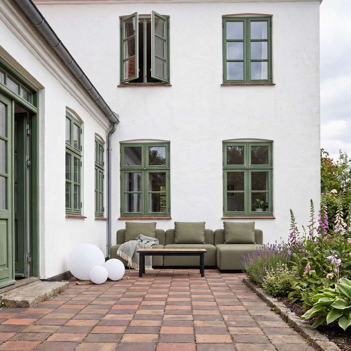 Olivegrønn tre seters loungesofa på en sjarmerende gårdsplass med hage og blomster, omgitt av hvite husvegger og grønne vinduskarmer