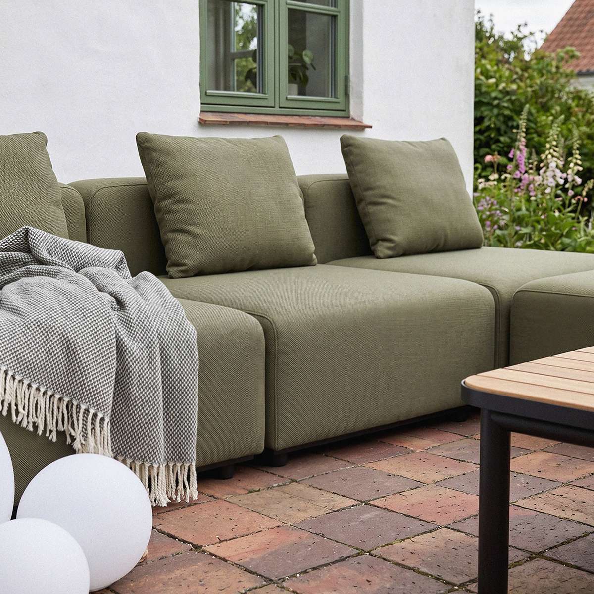 Olivengrønn modulbasert utesofa fra sackit med myke puter og pledd på en lun terrasse i en grønn hage