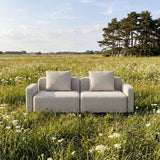 Lys grå toseters utesofa fra sackit i blomstrende eng med myke puter og skandinavisk design omgitt av høyt gress og trær