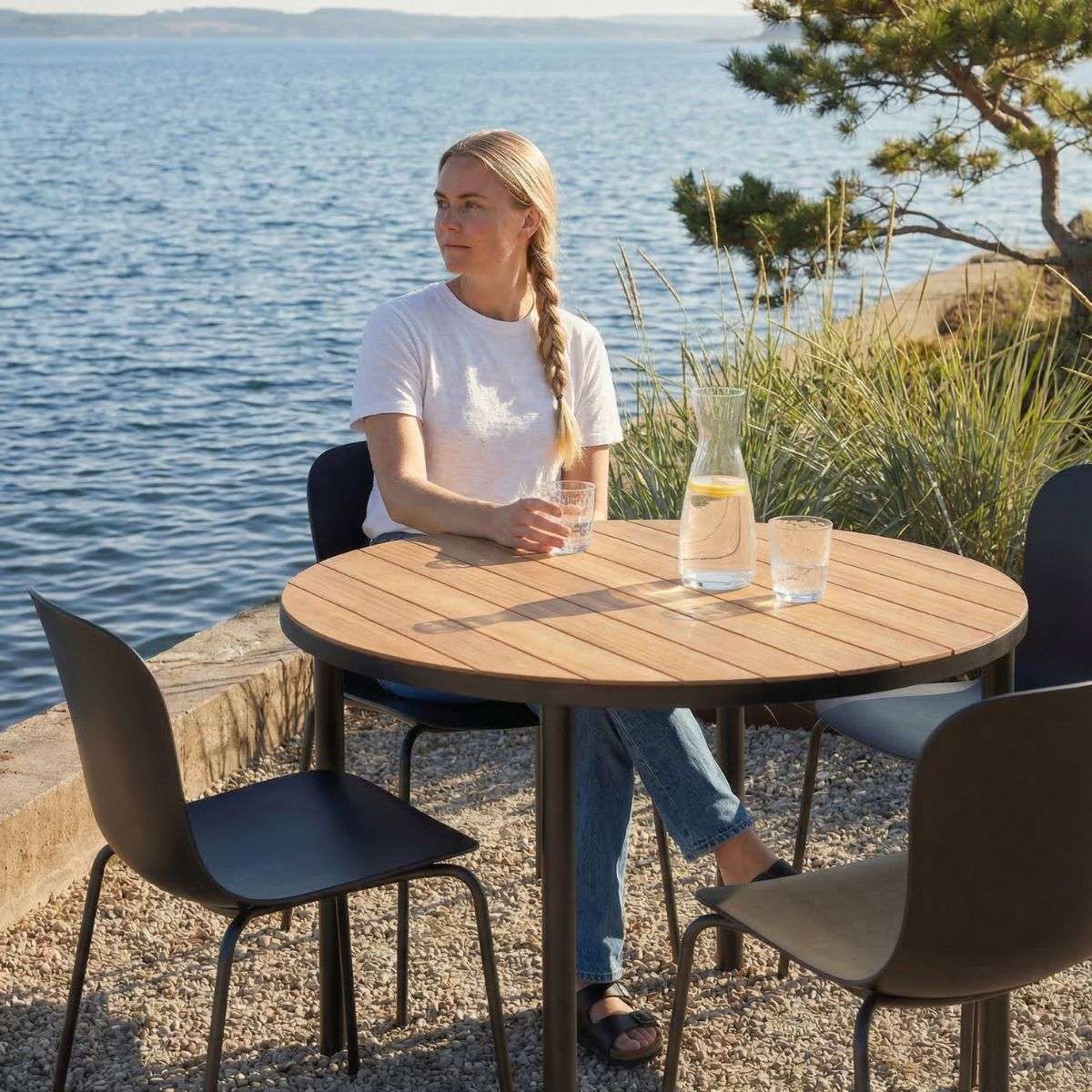 Rundt spisebord i teak med fire mørke hagestoler ved sjøen. Et nordisk utemiljø fra sackit med plass for måltider og hyggelige stunder.