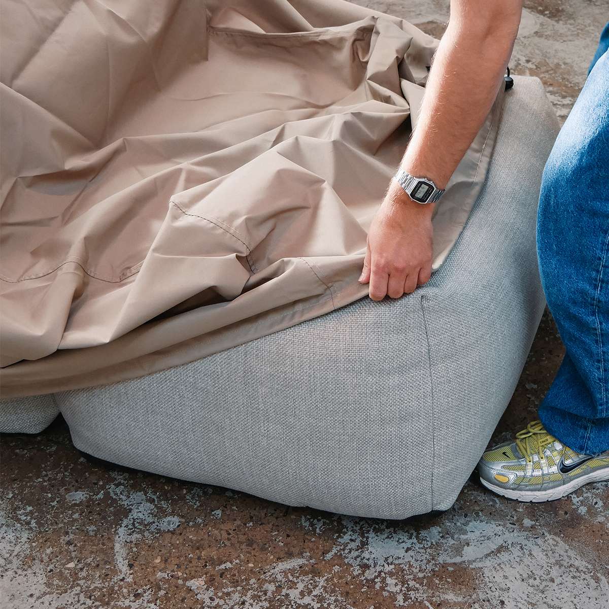 Person legger et sandfarget vind og vannavvisende trekk over en lys grå Soft Sofa fra sackit for beskyttelse mot vær og vind