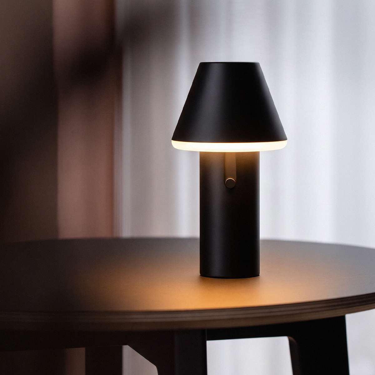 Sort trådløs bordlampe med konisk skjerm og mykt varmt lys på et mørkt bord. sackit Cone for innendørs og utendørs bruk