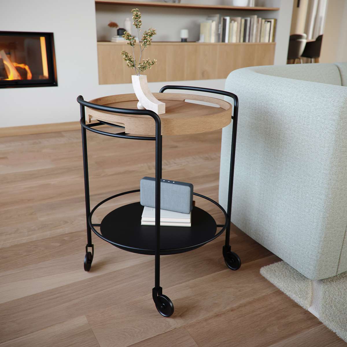 Rundt serveringsbrett i tre på et sort trillebord med hjul og nedre hylle ved siden av en lys sofa i nordisk stueinteriør