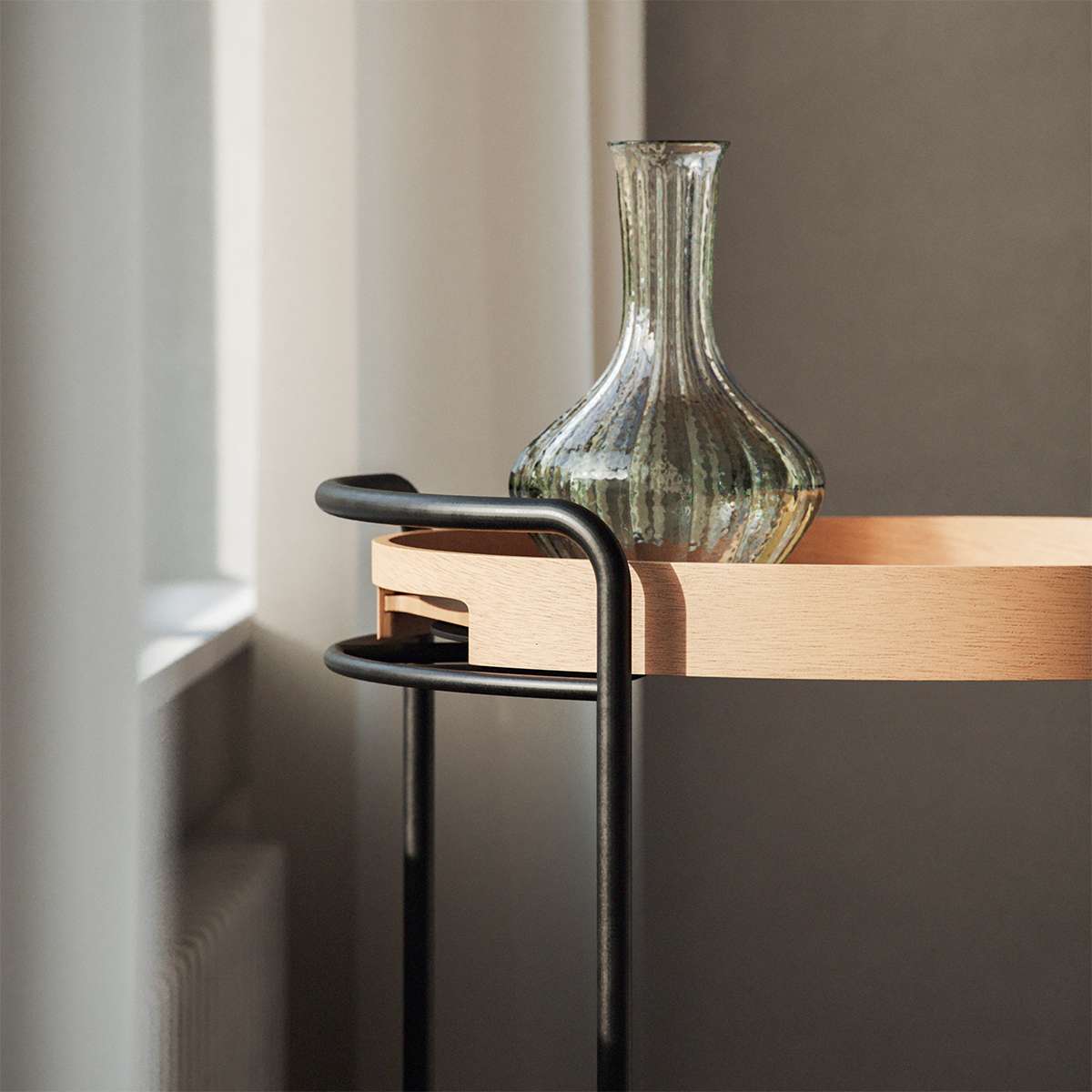 sackit serveringsbrett i lyst tre med rundt design på svart stativ og dekorativ glassvase i et lyst nordisk interiør