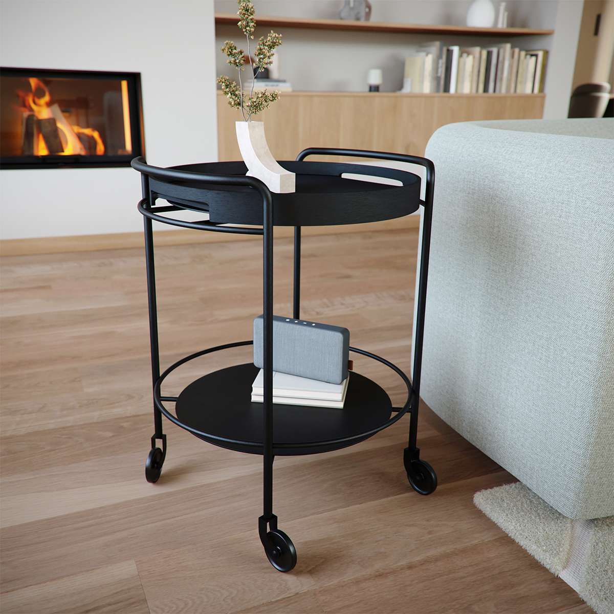 Sort serveringsvogn med to runde hyller og hjul i en nordisk stue med peis og lys sofa.