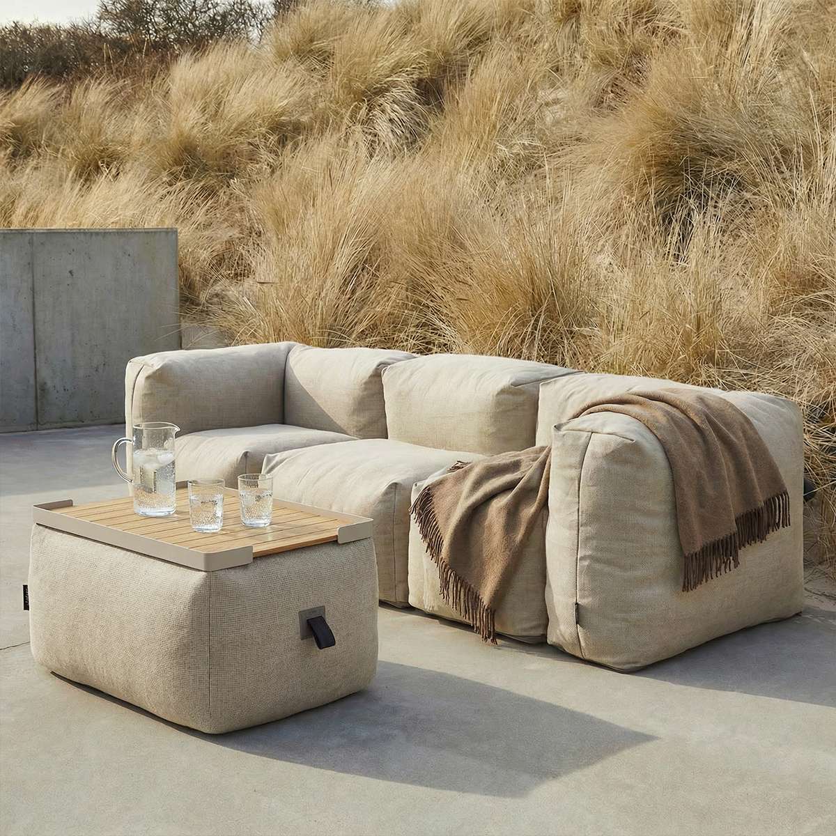 Beige utendørs pouf fra sackit som fotskammel og ekstra sitteplass foran en myk lounge sofa på en solrik terrasse ved sanddyner