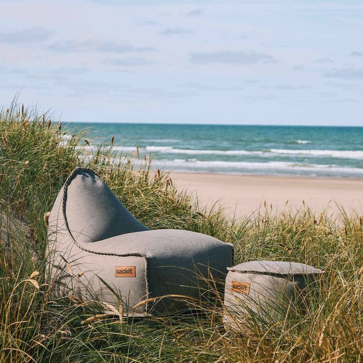 sackit Cobana loungestol og puff i sandfarget stoff omgitt av gressdyner med strand og hav i bakgrunnen