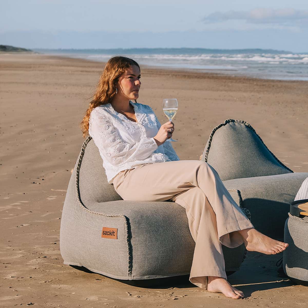 Kvinne sitter i en grå sackit Cobana loungestol på stranden med et glass vin ved sjøen en solrik dag