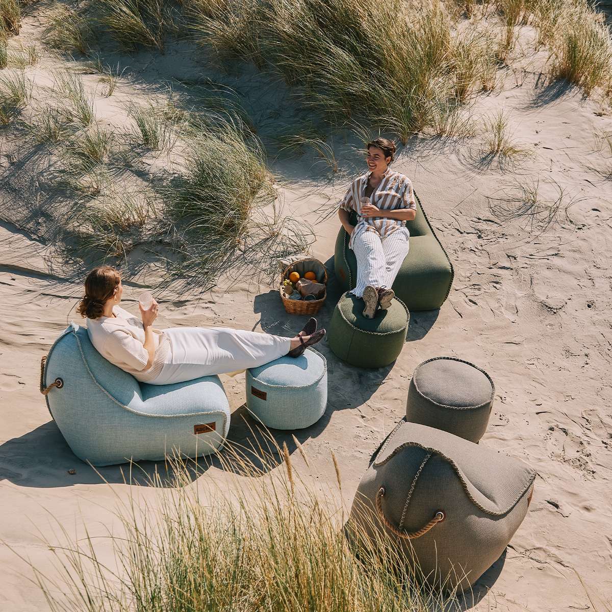 To personer slapper av i værbestandige saccosekker og puffer fra sackit på en sandstrand omgitt av marehalm og kystnatur