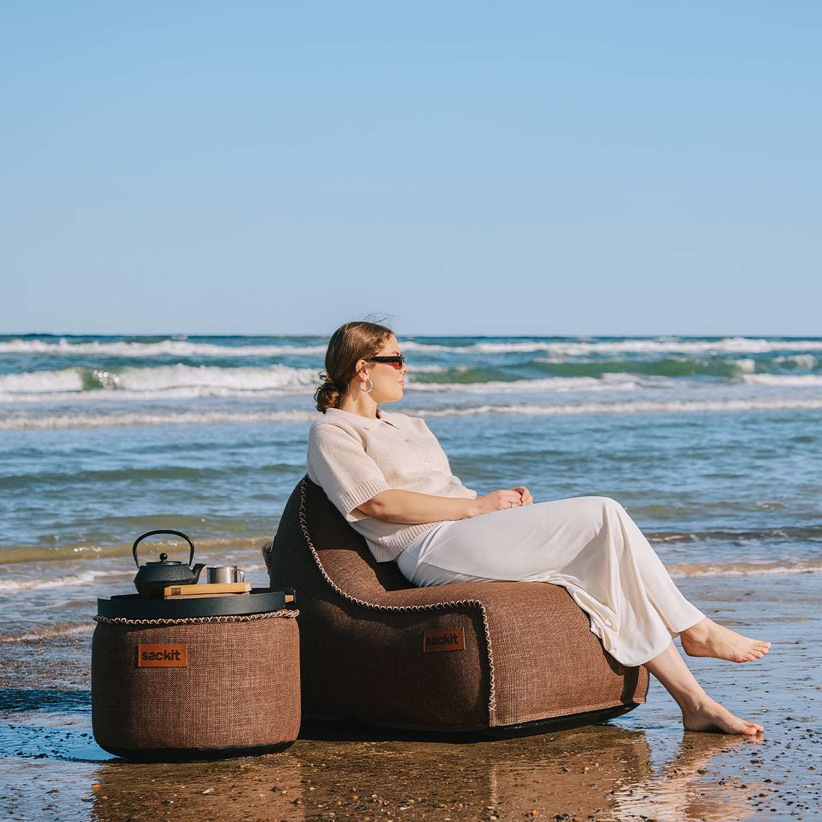 sackit Cobana loungestol og puff i brunt stoff på stranden ved havet mens en person slapper av i stolen