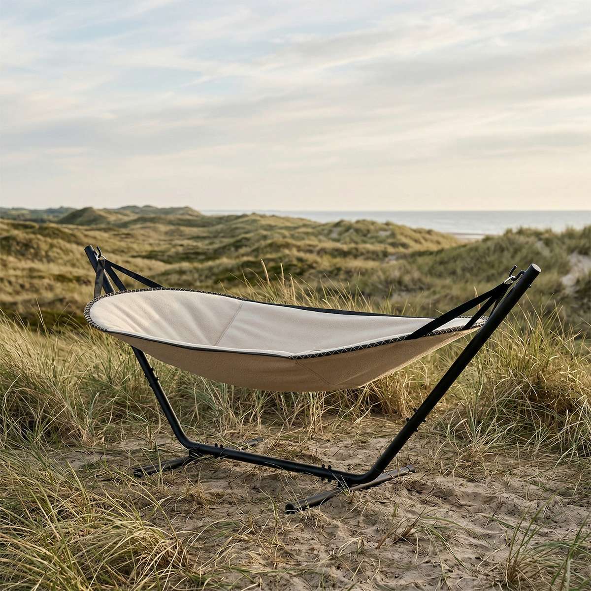 sackit Cobana hengekøye med svart stativ og mykt lyst tekstil plassert i sanddynene ved havet for avslappende utendørs komfort