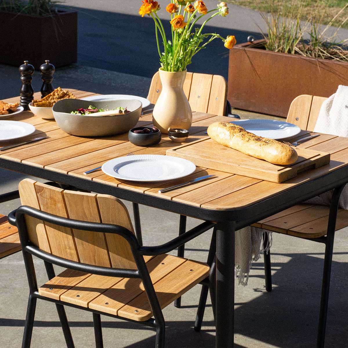 Kirra hagestol i teak og sort aluminium rundt et dekket hagebord med brød blomster og servise fra sackit