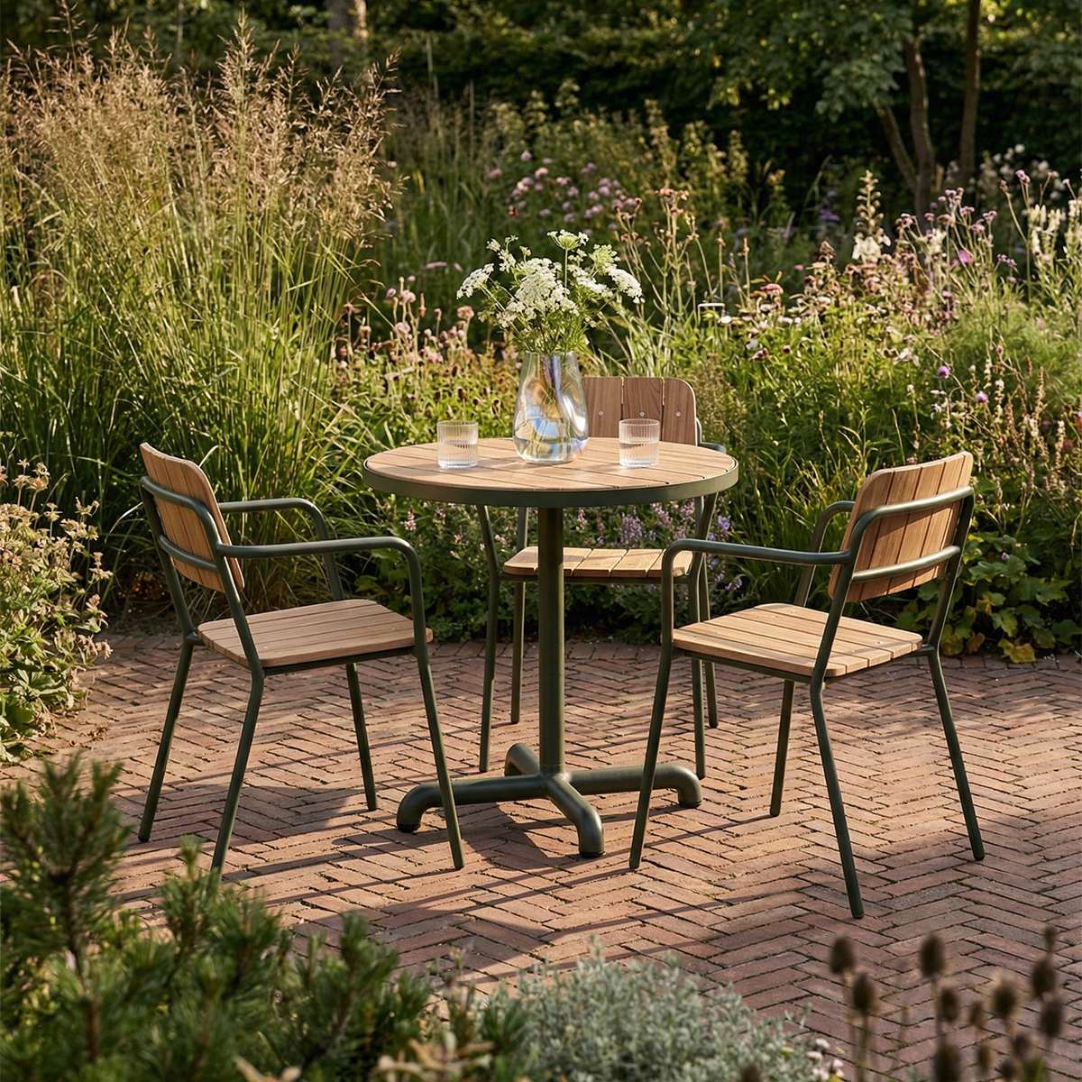Tre Kirra hagestoler i teak og mørkt pulverlakkert aluminium rundt et rundt hagebord på en solrik mursteinsterrasse omgitt av frodig beplantning