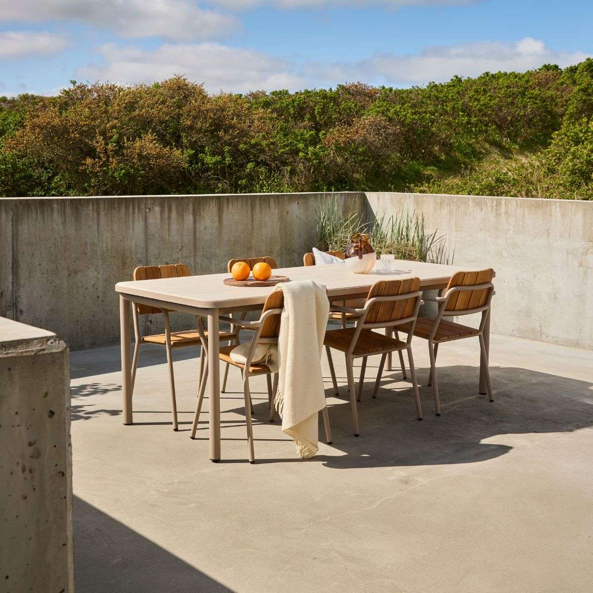 Utendørs spisegruppe med lyst bord og seks trestoler på en solrik betongterrasse omgitt av grøntområder. Designet av sackit for nordisk utebruk.