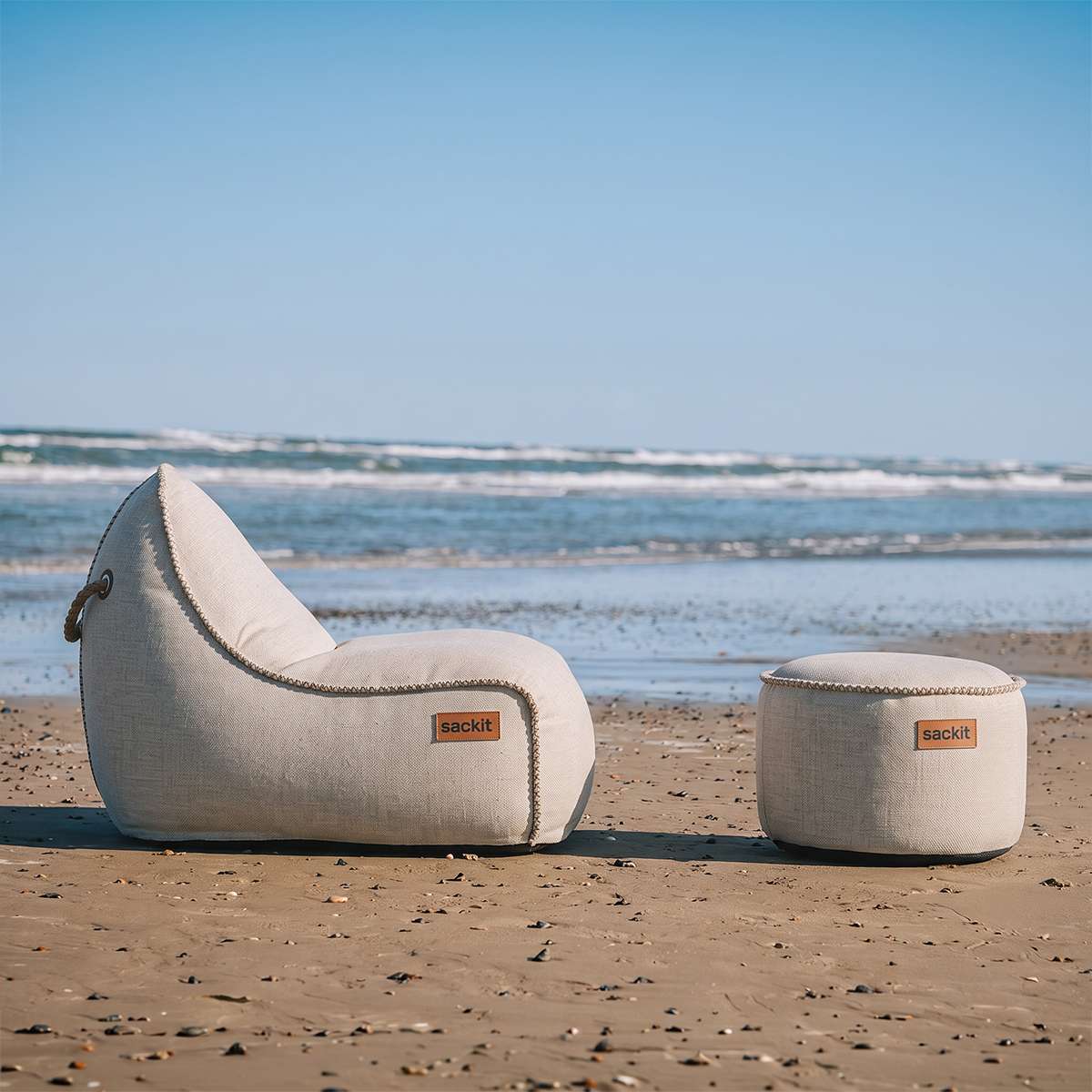 sackit Cobana loungestol og puff i lyst tekstil på en sandstrand med havet i bakgrunnen, designet for avslapping utendørs