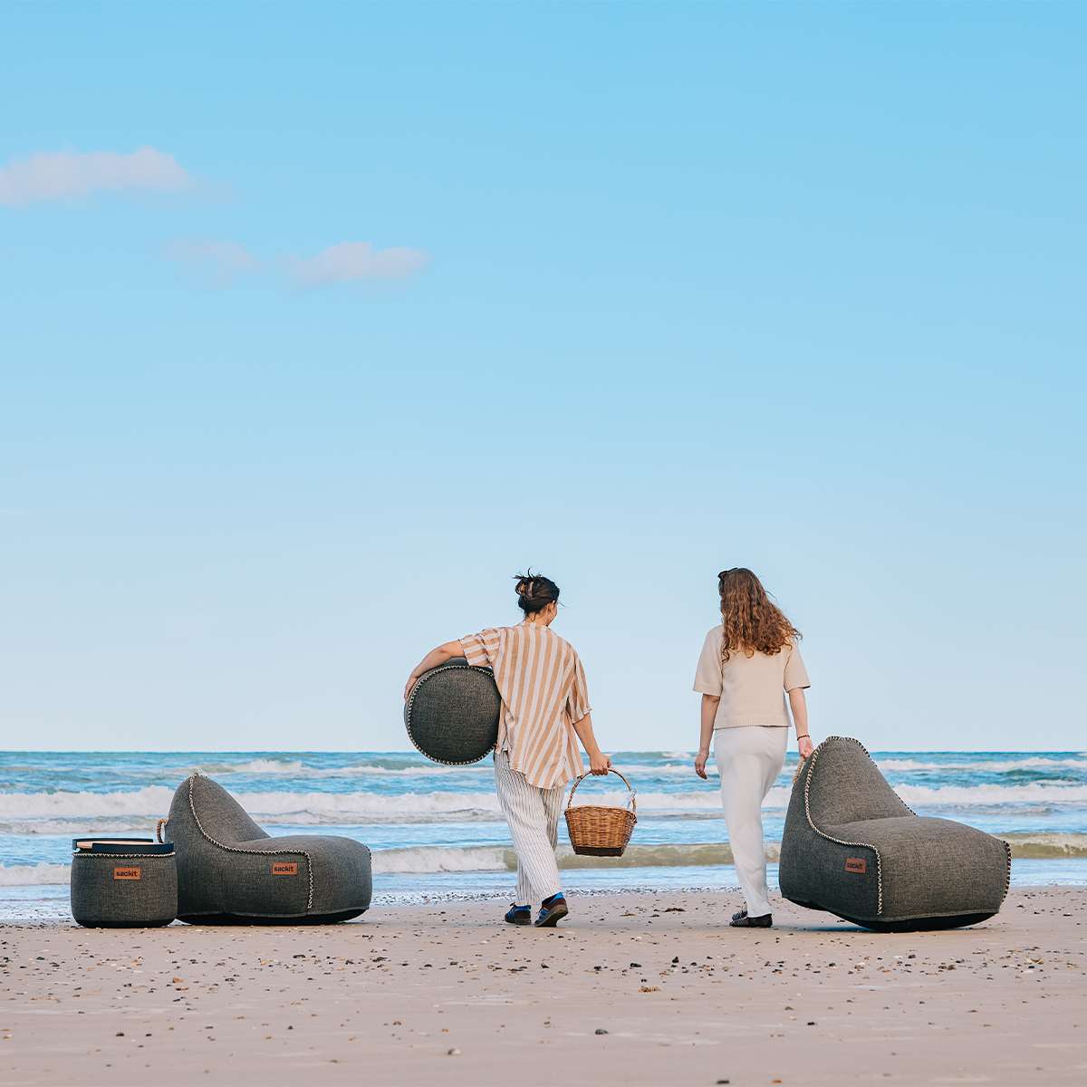To mørke loungestoler med puffer fra sackit på stranden, mens to personer går langs vannkanten i solfylt nordisk sommerlandskap