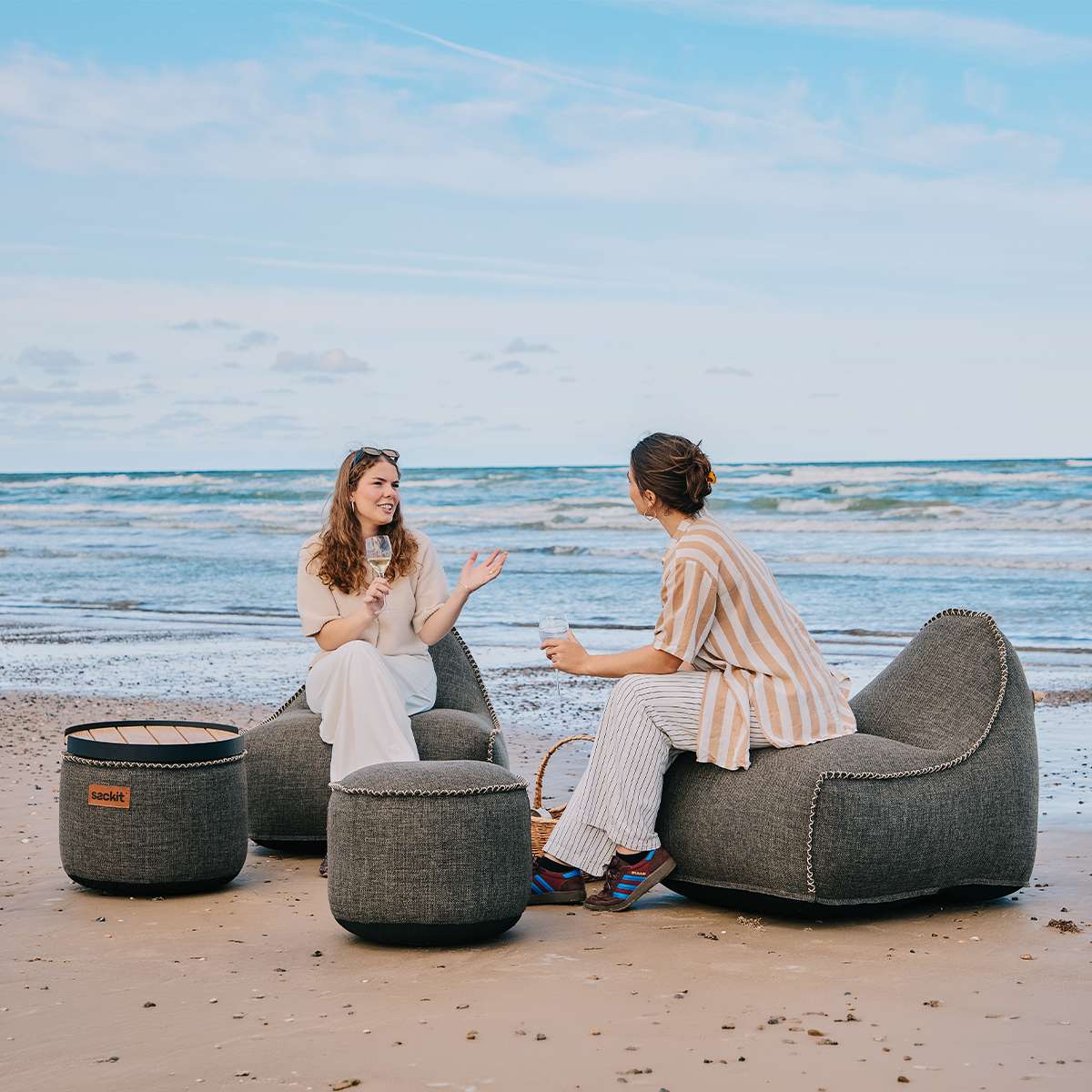 To kvinner slapper av på stranden i værbestandige Cobana loungestoler og puff fra sackit med havet i bakgrunnen, designet for nordisk utendørsbruk