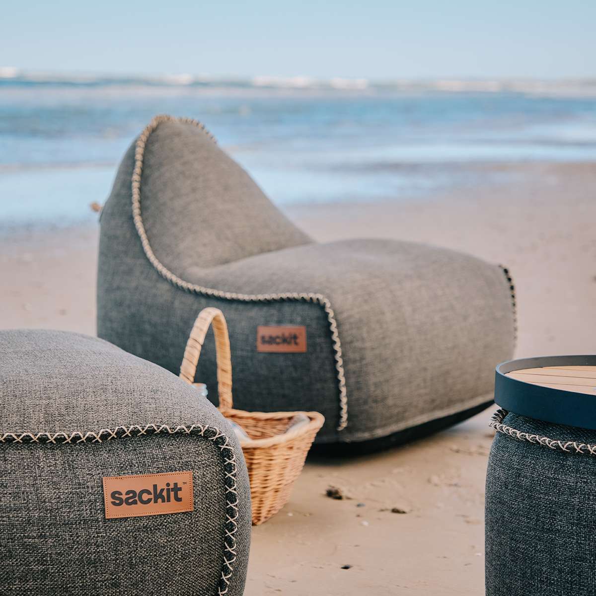 Grå utendørs loungestol og puff fra sackit på en sandstrand, med kurv og rundt sidebord i forgrunnen og havet i bakgrunnen