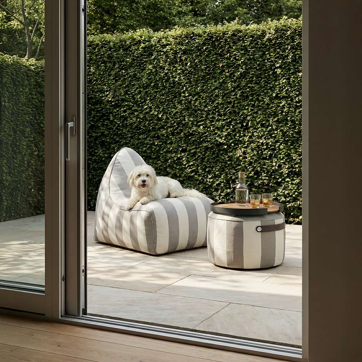 Stripete sackit loungestol utendørs på terrasse med en liten hund og rundt sidebord foran en grønn hekksett.