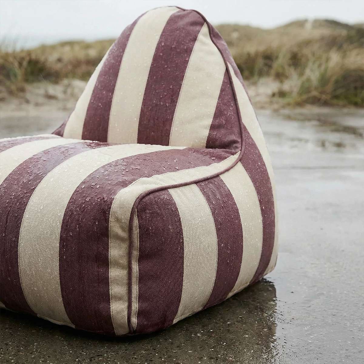 Stripete utendørs loungestol fra sackit i burgunder og kremfarget tekstil på en våt strand med sanddyner i bakgrunnen