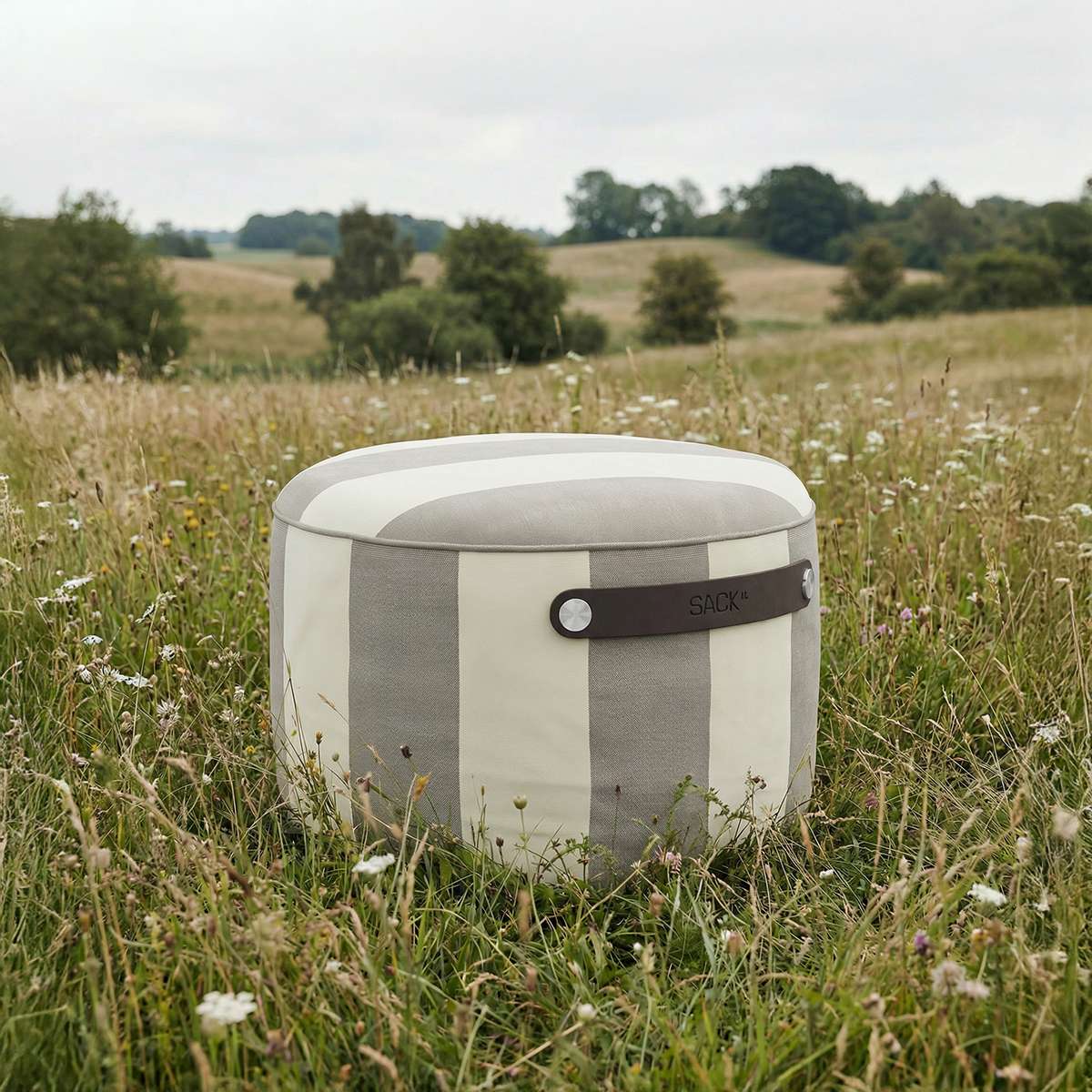 Rund utendørs puff fra sackit i lyse tekstilflater plassert i en blomstrende eng med åkerlandskap og trær i bakgrunnen