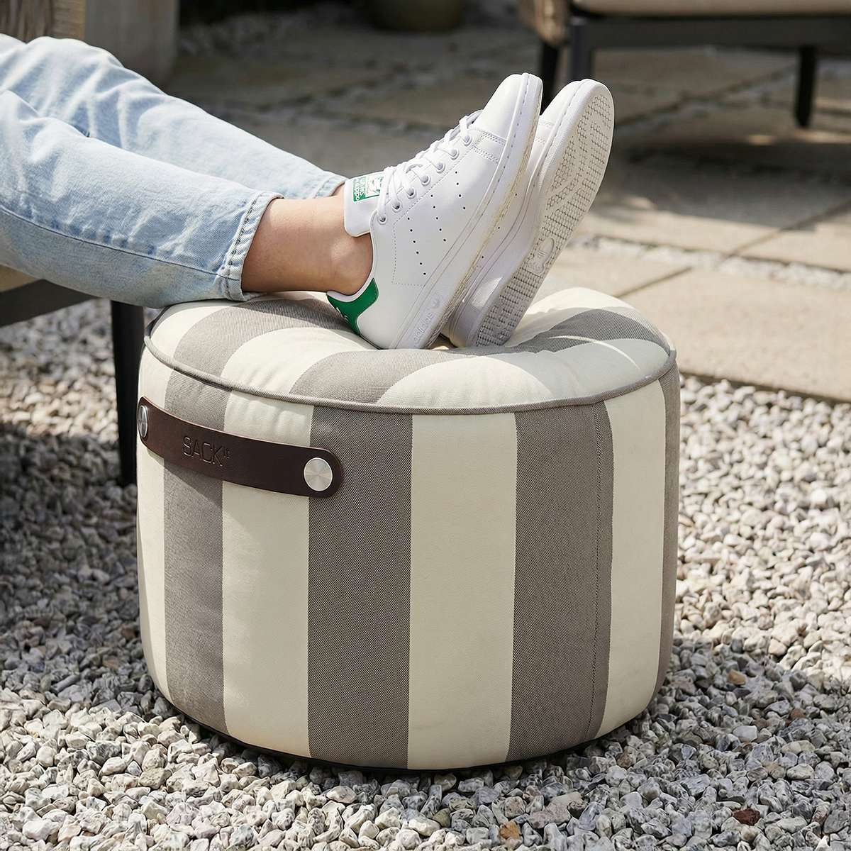Stripete utendørs puff fra sackit i grått og kremhvitt med avrundet form og føtter i hvite joggesko på en solrik terrasse med singelgrus